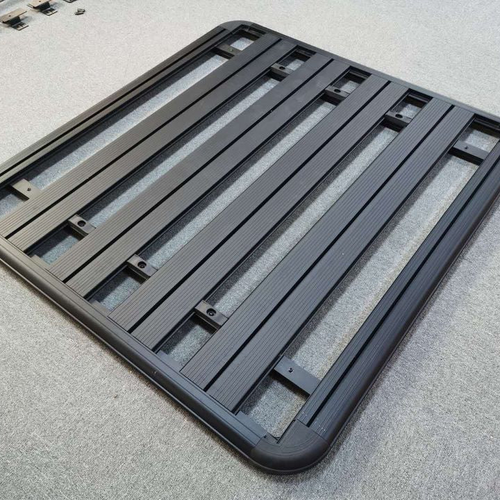 Black aluminum platform-style roof rack, composed of a perimeter frame with rounded corners and several horizontal slats with integrated T-slots for mounting accessories.