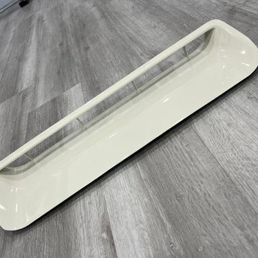 A long, rectangular, beige or off-white plastic grille or vent insert, likely for an automobile dashboard, interior, or exterior body panel. It features a wide, recessed opening down the center, which is spanned by a thin horizontal bar supported by several small vertical struts. The part is sitting on a gray, wood-look plank floor.