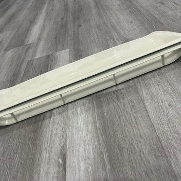 A long, narrow, beige or off-white air vent cover or deflector, likely made of plastic, resting on a gray, wood-look plank floor. The part has a slightly curved profile with rounded ends and features a recessed central channel with several vertical supporting ribs visible underneath.