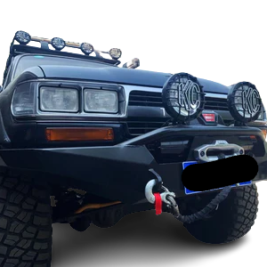 Close-up of the front of a dark colored, off-road equipped vehicle, likely a Toyota Land Cruiser 80-Series. It features a heavy-duty black steel bumper with a visible winch opening (license plate area redacted), two prominent round KC auxiliary lights with black grilles, a large tow hook with a red recovery strap, and a set of smaller round LED lights mounted on a roof rack visible above the windshield. The background is a checkered transparency pattern.