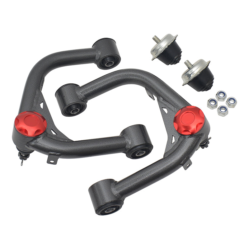 Two gray aftermarket upper control arms with a slightly metallic finish, featuring red adjustable caps on the ball joint housings. The control arms are angled and crossed, positioned near two separate black and silver polyurethane or rubber bushings, and a small cluster of four silver nuts with blue locking inserts. The parts are for a vehicle's suspension system.