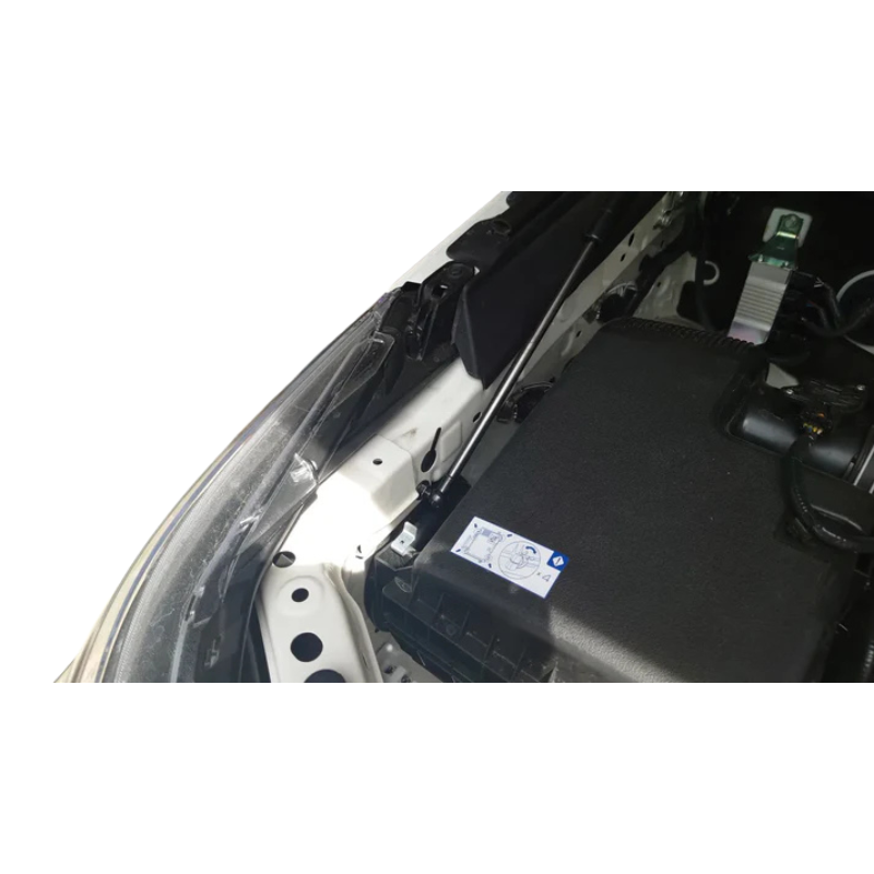 A close-up photograph showing the installation of a hydraulic hood strut (gas spring) in the engine bay of a white vehicle. The silver strut rod is mounted low on the fender/chassis structure next to the headlight assembly and above a large black component cover (likely the airbox or a fuse box). The white paint of the chassis and the black plastic components contrast with the bright lighting.