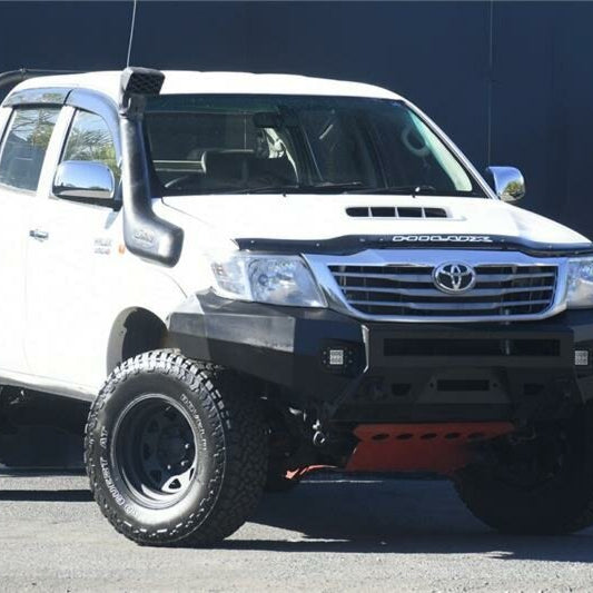A close-up, angled front view of a white Toyota Hilux pickup truck modified for off-roading. The vehicle is fitted with a black snorkel on the driver's side, a heavy-duty black steel off-road bumper, and a red bash plate/skid plate visible underneath. The truck also has large, black off-road wheels and a black hood deflector.