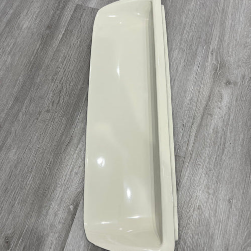 A side view of a long, rectangular, beige or off-white plastic cover or trim piece, possibly for an appliance or vehicle interior. It has a smooth, curved front profile and is resting vertically on a gray, wood-look plank floor.