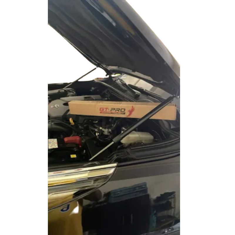 A dark-colored vehicle (possibly black) is shown with its hood open, revealing the engine bay. A pair of hydraulic hood struts are already installed, holding the hood up. Resting across the engine is a long, brown cardboard box with red and black text, featuring the "GT-PRO" logo, which is the packaging for the hood strut kit. The image appears to be taken after the product was installed.