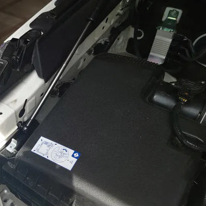 A close-up view of a hydraulic hood strut (gas spring) installed in the engine bay of a white vehicle. The silver rod of the strut is clearly visible, extending diagonally upwards from a mounting point on the white chassis/fender. The strut passes over a large, black textured plastic component, likely the air box, which has a small blue and white instructional sticker on it. The image focuses on the strut installation location.