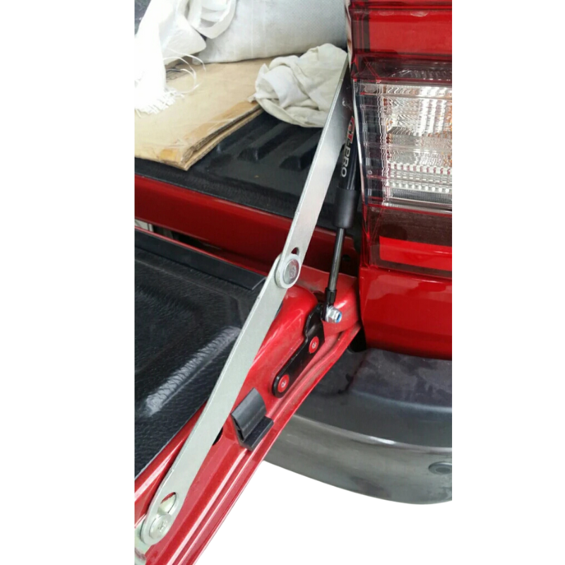 A close-up shot of a tailgate damper/assist strut installation on a red pickup truck. A silver metal bracket is visible, which appears to be a limiting or mounting arm, connected to a black hydraulic strut labeled "GT-PRO." The strut is mounted to the red body/tailgate near the taillight assembly and a black plastic bumper or trim piece. The truck bed has a black textured liner.