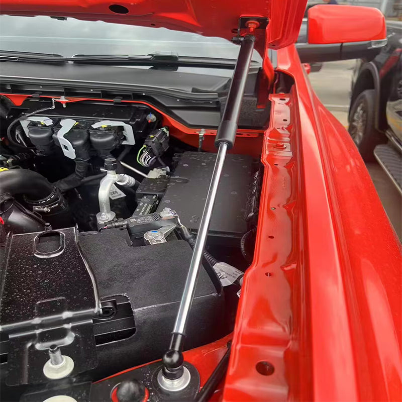 A close-up image showing a black and silver gas spring strut installed on a bright red vehicle, holding the hood open. The strut extends from a ball stud mount near the fender area of the engine bay up to a mounting point on the underside of the hood. The vehicle's engine and various components are visible in the background, which is partially obscured by the open hood. The strut is fully extended, indicating the hood is up.