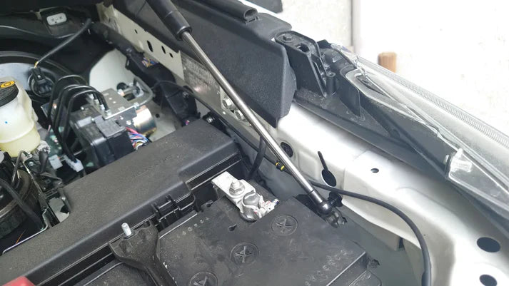 A close-up image showing the lower mounting point of a hydraulic hood strut (gas spring) in a vehicle's engine bay. The strut's silver rod extends diagonally from the chassis mount. The mount is installed near the vehicle's battery, which is a large black box, and adjacent to the headlight assembly and other engine components like the ABS module. The view is focused on the strut installation.