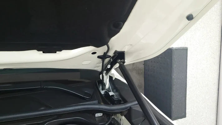 A close-up view of a hydraulic hood strut (gas spring) installed on a white vehicle. The strut connects the underside of the hood (which is open) to the engine bay area near the hinge. The mounting bracket and the black piston of the strut are clearly visible against the white paint and black insulated hood liner.