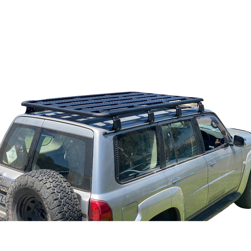 A rear three-quarter view of a silver 4x4 SUV, likely a Nissan Patrol, featuring a full-length, black basket-style roof rack platform mounted over the entire passenger compartment. The rack has an outer perimeter rail and multiple parallel internal slats. The rack is secured to the roof via black mounting feet. The vehicle also has a spare tire mounted on the rear door. The background is isolated in white.