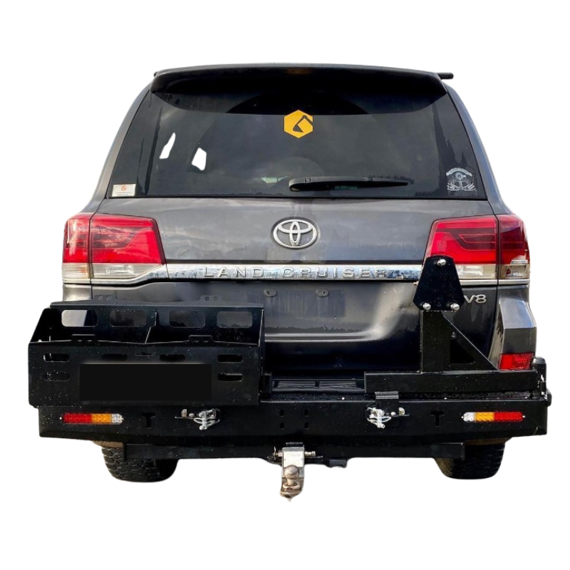 A rear view of a dark gray Toyota Land Cruiser 200 Series SUV, heavily modified with a heavy-duty, black steel rear bar and swing-out carrier system.
The rear bar is mounted below the tailgate and features:
A swing-out jerry can/storage carrier on the left side, with multiple compartments visible.
A swing-out spare tire carrier on the right side, with a mounting plate visible.
Integrated lights (reverse/indicators) on both ends of the bumper.
A tow hitch with a tow ball visible below the bumper.