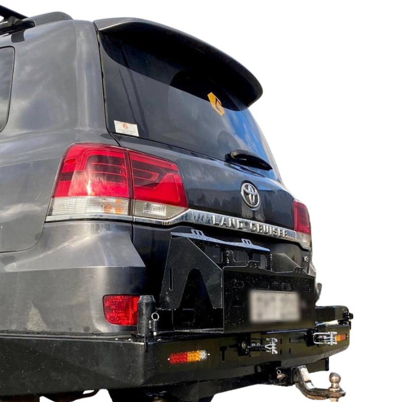 A close-up, rear quarter-view image of a dark gray Toyota Land Cruiser 200 Series SUV, showing the heavy-duty, black steel rear bumper with a swing-out carrier attached.
The main focus is the rear bumper which has integrated amber and red lights on the corner. The swing-out carrier is shown in its closed position, with the black steel frame visible directly beneath the vehicle's tailgate, likely for mounting a spare tire or jerry cans. A tow hitch with a tow ball is clearly visible.