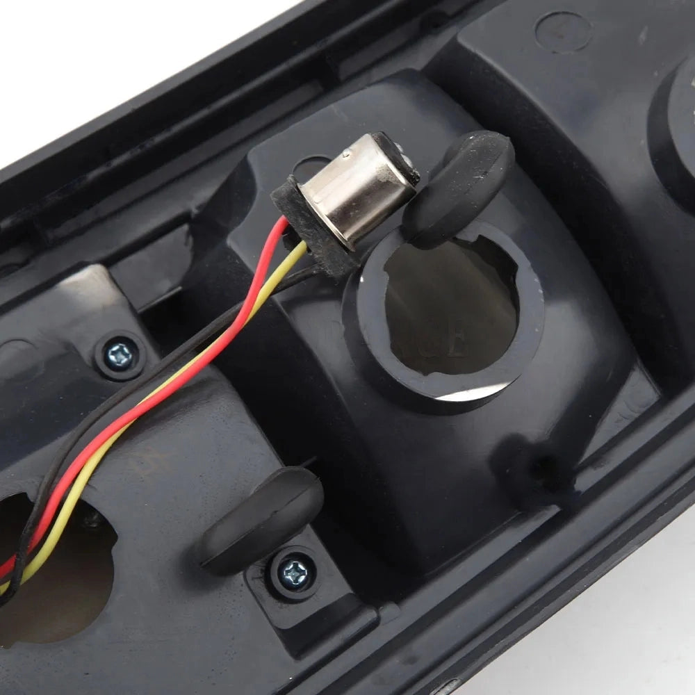 A close-up view of the lower backside of a black vehicle taillight assembly, showing a small metal bulb socket wired with red, black, and yellow wires. The socket is partially inserted into the plastic housing next to a larger, empty circular opening, with two rubber grommets also visible.