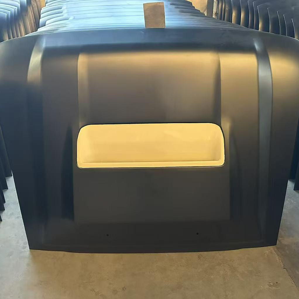 A stack of dark-colored, possibly black, car hoods is shown in a storage or warehouse setting. The topmost hood, seen from the front, features a prominent rectangular hood scoop opening. The main surface of the hood is matte black, while the inside of the scoop is a contrasting light cream or beige color, indicating it may be the raw fiberglass or unpainted substrate. The other hoods are stacked closely behind it.