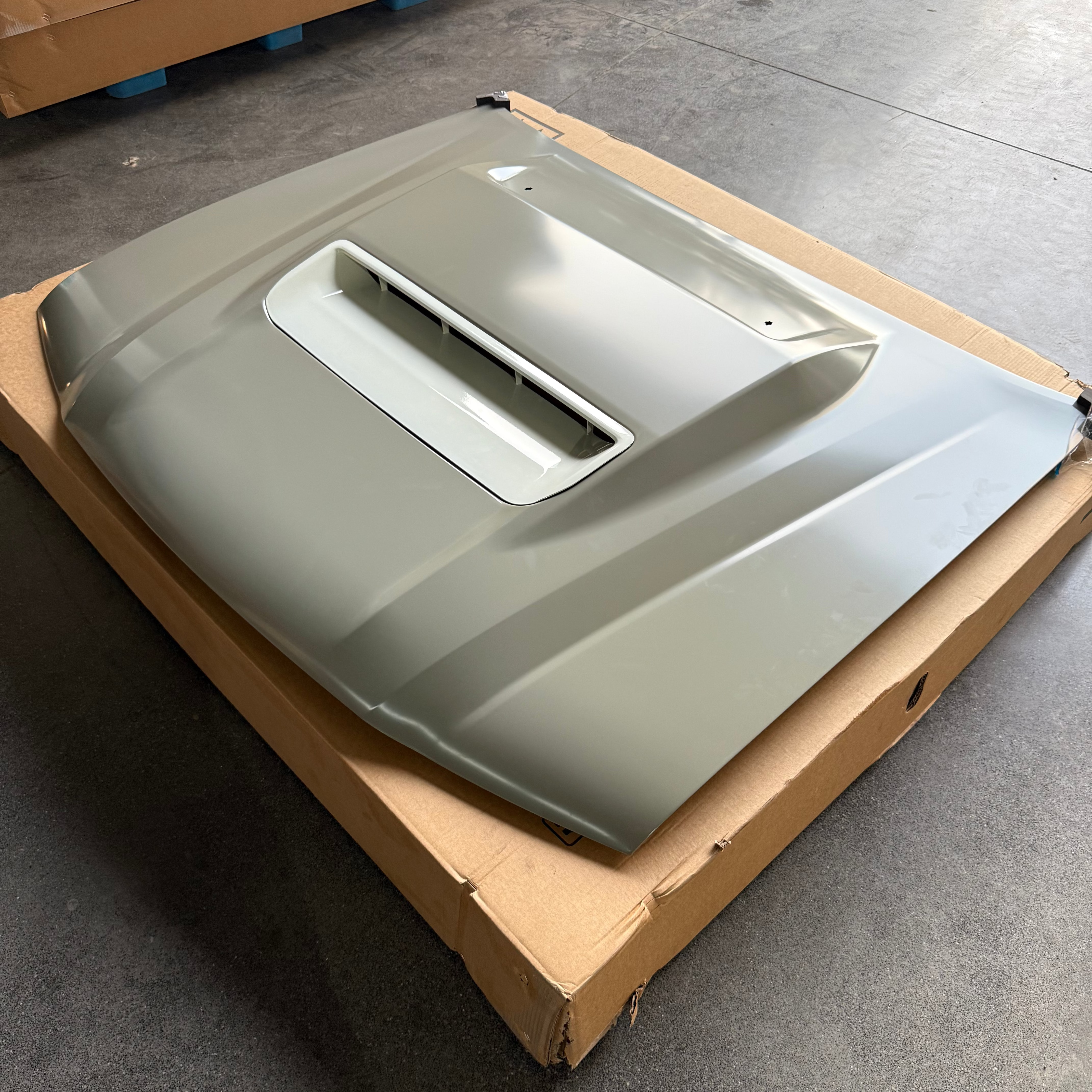 A close-up photograph of a car hood with a distinct hood scoop positioned centrally. The hood is painted a matte light gray or beige color and rests on top of a large, flattened cardboard box. The hood scoop insert appears to be a slightly lighter or unpainted material. The setting looks like a warehouse or garage floor.
