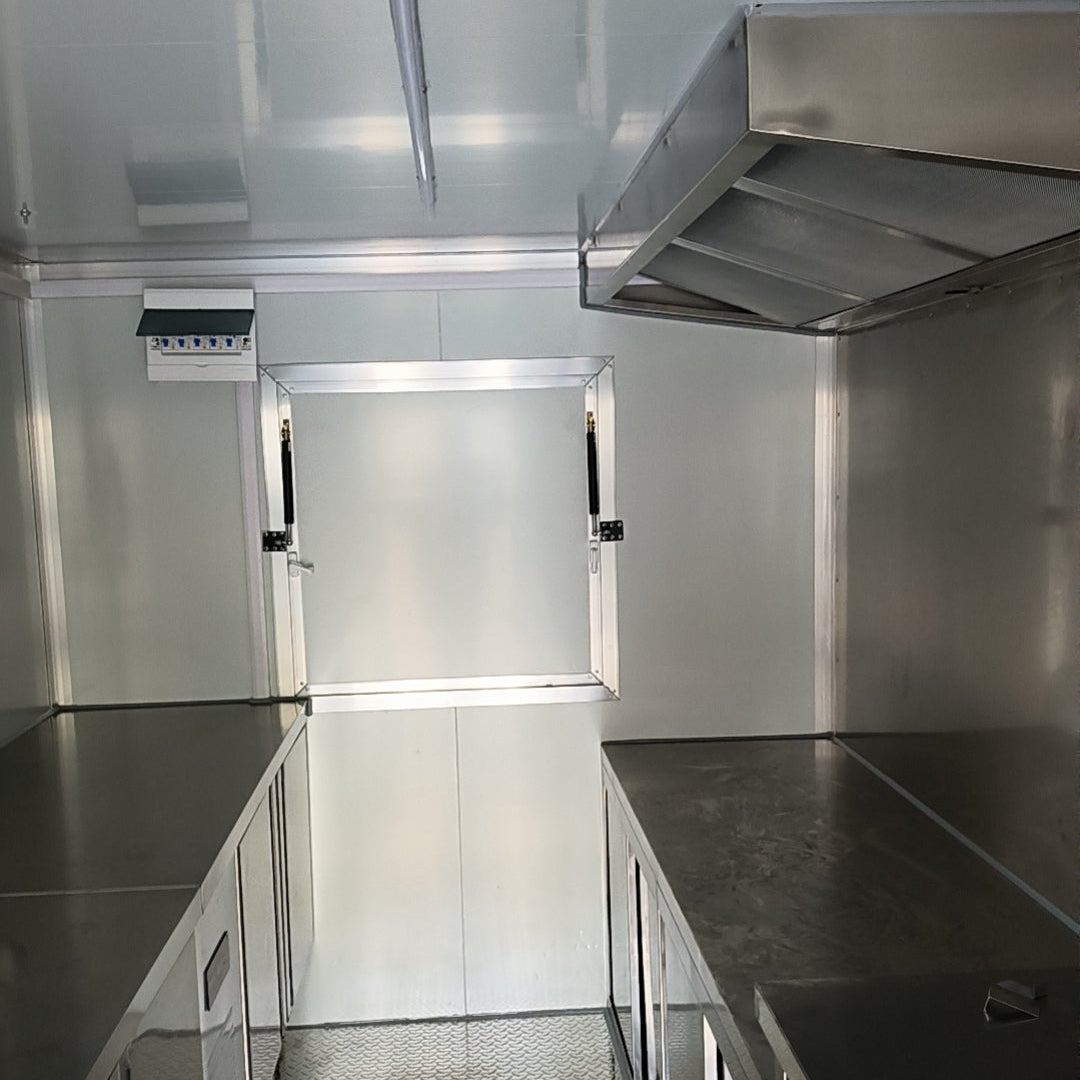 The interior of a clean, bright, commercial food trailer kitchen. The space features stainless steel countertops and lower cabinets on both sides, a large stainless steel ventilation hood on the right, and an open service window in the back wall. A small electrical breaker panel is mounted on the wall next to the window, and the floor has a textured, slip-resistant surface.