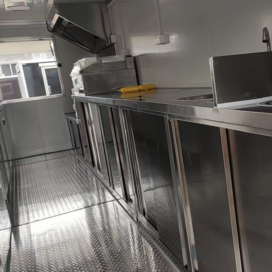 A clean, newly installed professional kitchen interior, likely within a food truck or mobile catering trailer. The floor is made of reflective diamond plate aluminum. Along the right side, there is a long stainless steel countertop with multiple under-counter storage cabinets and a sink area. Above the counter, there's a stainless steel exhaust hood and white metal walls with electrical outlets. A small window is visible on the far left wall, looking out onto a street or building exterior.