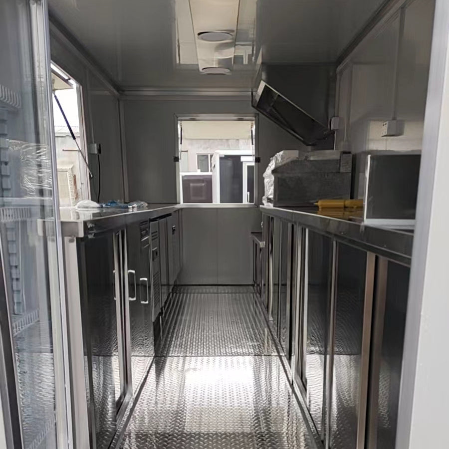 A perspective view looking down the aisle of a stainless steel food trailer kitchen. The interior features reflective stainless steel countertops and lower cabinets lining both sides, and a diamond-plate metal floor. There is a service window open at the far end, showing a glimpse of the outside. A stainless steel ventilation hood and some equipment are visible on the right countertop.