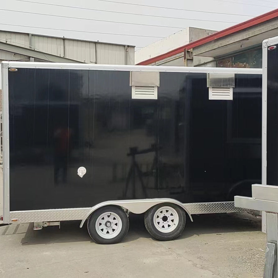A horizontal photo capturing the side profile of a long, black enclosed trailer, likely a concession or food service trailer. The side panel is smooth and black with two rectangular, louvered silver vents near the top, indicating ventilation for cooking equipment inside. The lower portion of the trailer features a silver diamond plate trim. The trailer is supported by tandem axles with two white wheels featuring five triangular spokes. The background includes parts of industrial buildings with metal siding