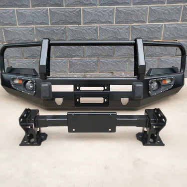 A heavy-duty, black metal bull bar or replacement bumper assembly for a truck or off-road vehicle. The main bumper unit features a wrap-around tubular grille guard, integrated fog lights and turn signals, and a central opening for a winch. Below it is a separate black metal mounting bracket or tow bar. The parts are resting in front of a dark gray brick wall.