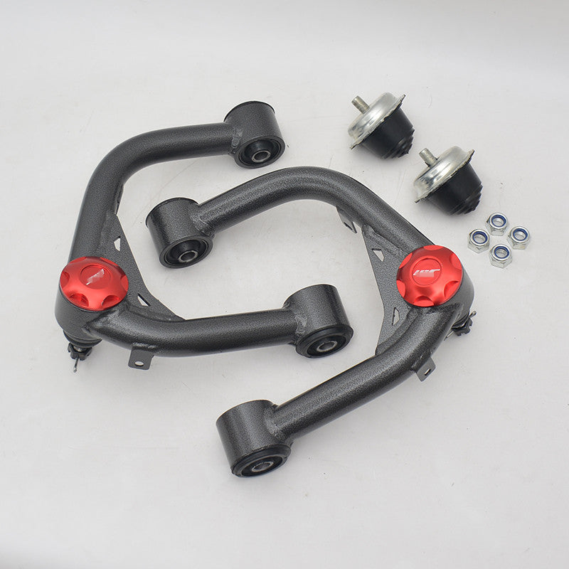 Overhead shot of a pair of aftermarket upper control arms (UCAs) for a vehicle suspension, laid out on a white background. The UCAs have a dark gray, textured finish and feature bright red aluminum dust caps on the ball joint housings. Also included in the kit are two metal and rubber suspension mounts and a set of four locking nuts.
