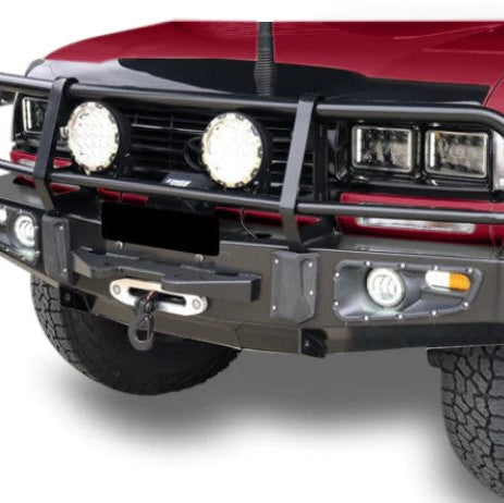 A close-up view of the front of a red vehicle (likely an SUV or truck) heavily equipped with a black steel off-road bull bar. The bar features a winch roller fairlead and hook at the center, two large circular auxiliary spotlights mounted above the bumper, and integrated fog lights and turn signals in the wings. The vehicle has square headlights and large off-road tires visible at the bottom.