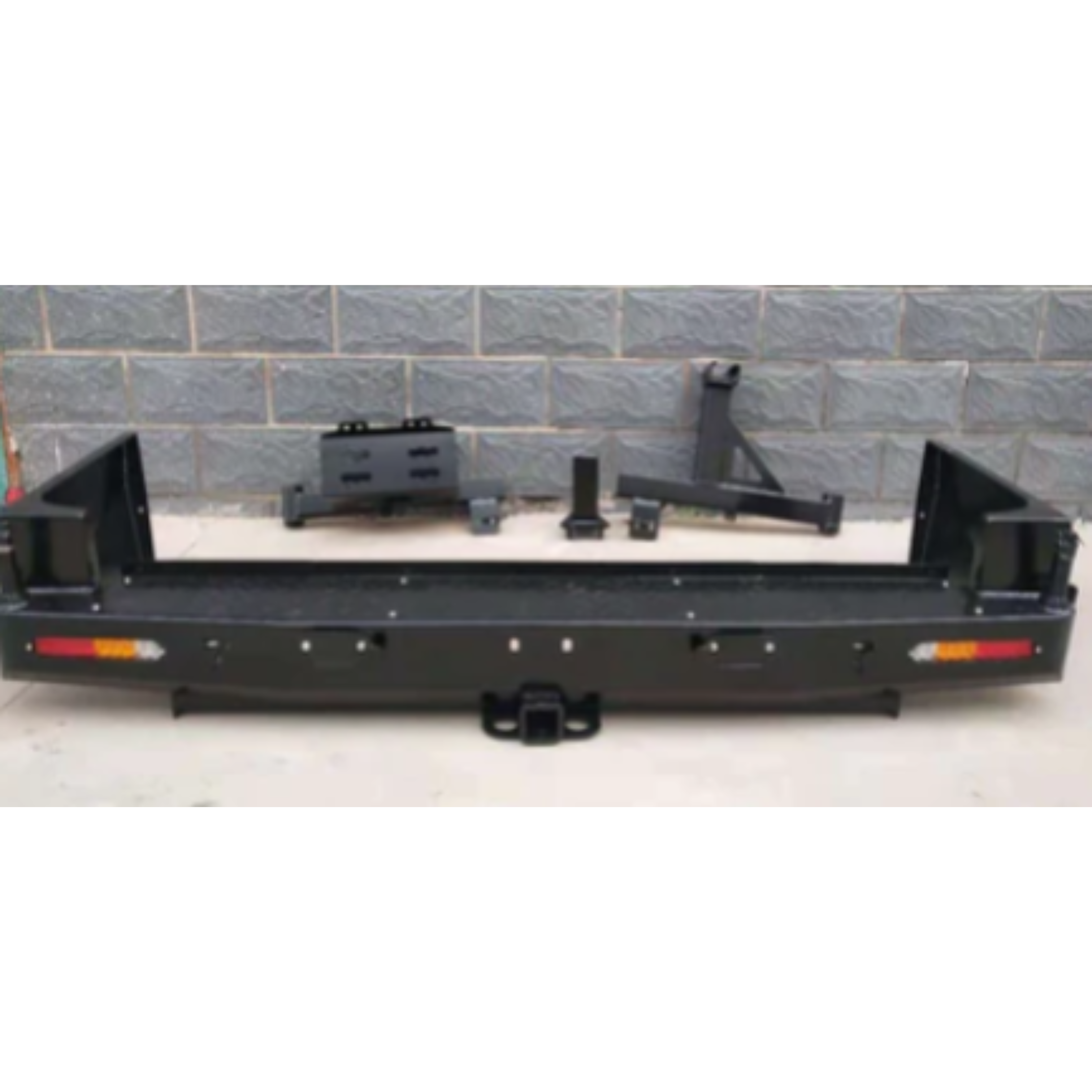A wide, straight-on product shot of a heavy-duty, flat black steel rear bumper kit for a 4x4 or off-road vehicle.
The main bumper piece includes:
A central receiver hitch with a tow ball mount attached.
Integrated amber and red lights on the far left and right ends.
Deep recesses on the sides, likely for mounting to the vehicle frame.
The additional components are laid out above the bumper against a gray brick wall background. These pieces include a spare tire carrier mount (a triangular, angled piece)
