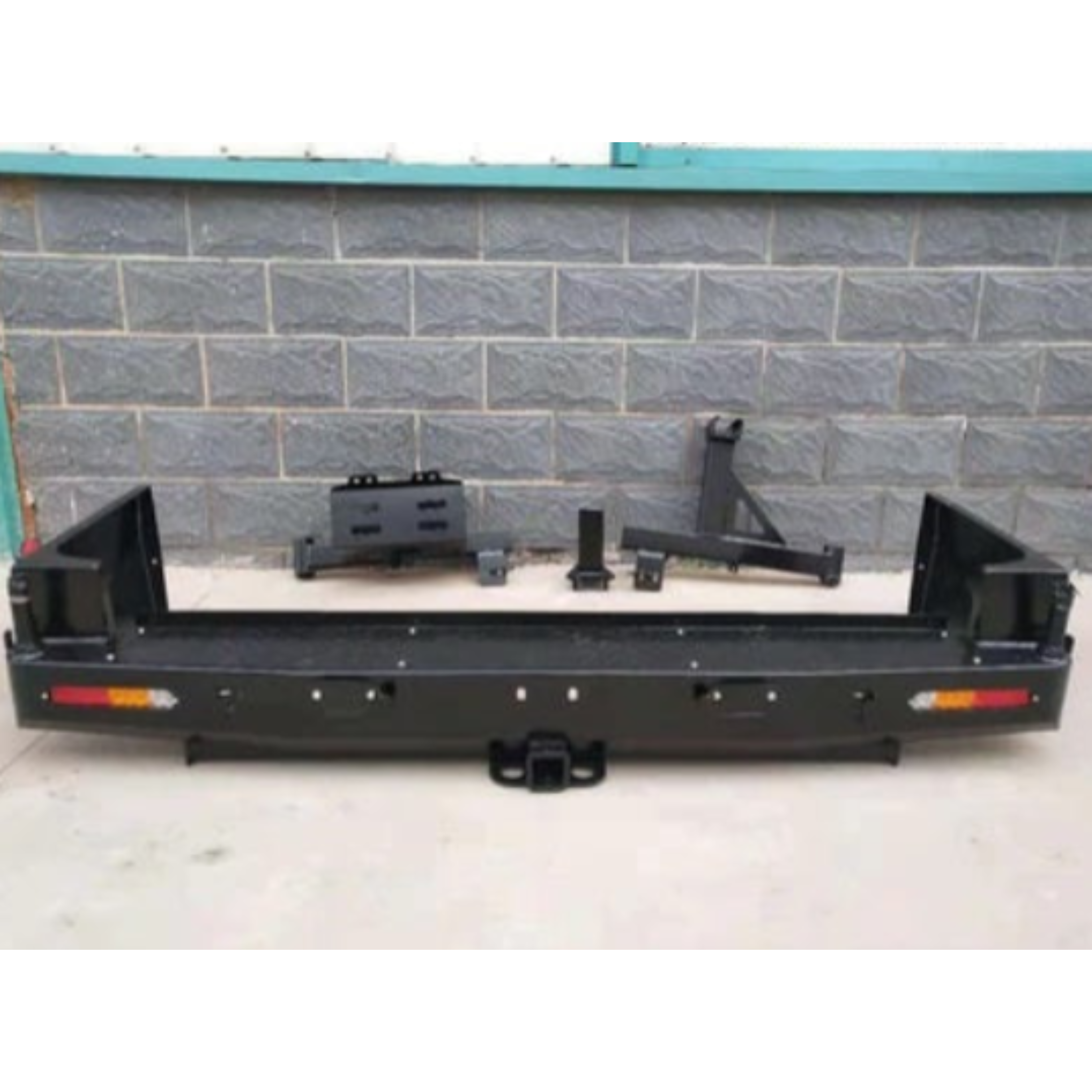 A heavy-duty, black steel rear bumper kit, likely for an off-road vehicle or truck, resting on a concrete floor in front of a dark gray cinder block wall. The main bumper assembly includes integrated red and amber lights on both ends and a central receiver hitch. Several separate accessory pieces, including mounting brackets and components for what looks like a spare tire carrier, are placed behind the main bumper.