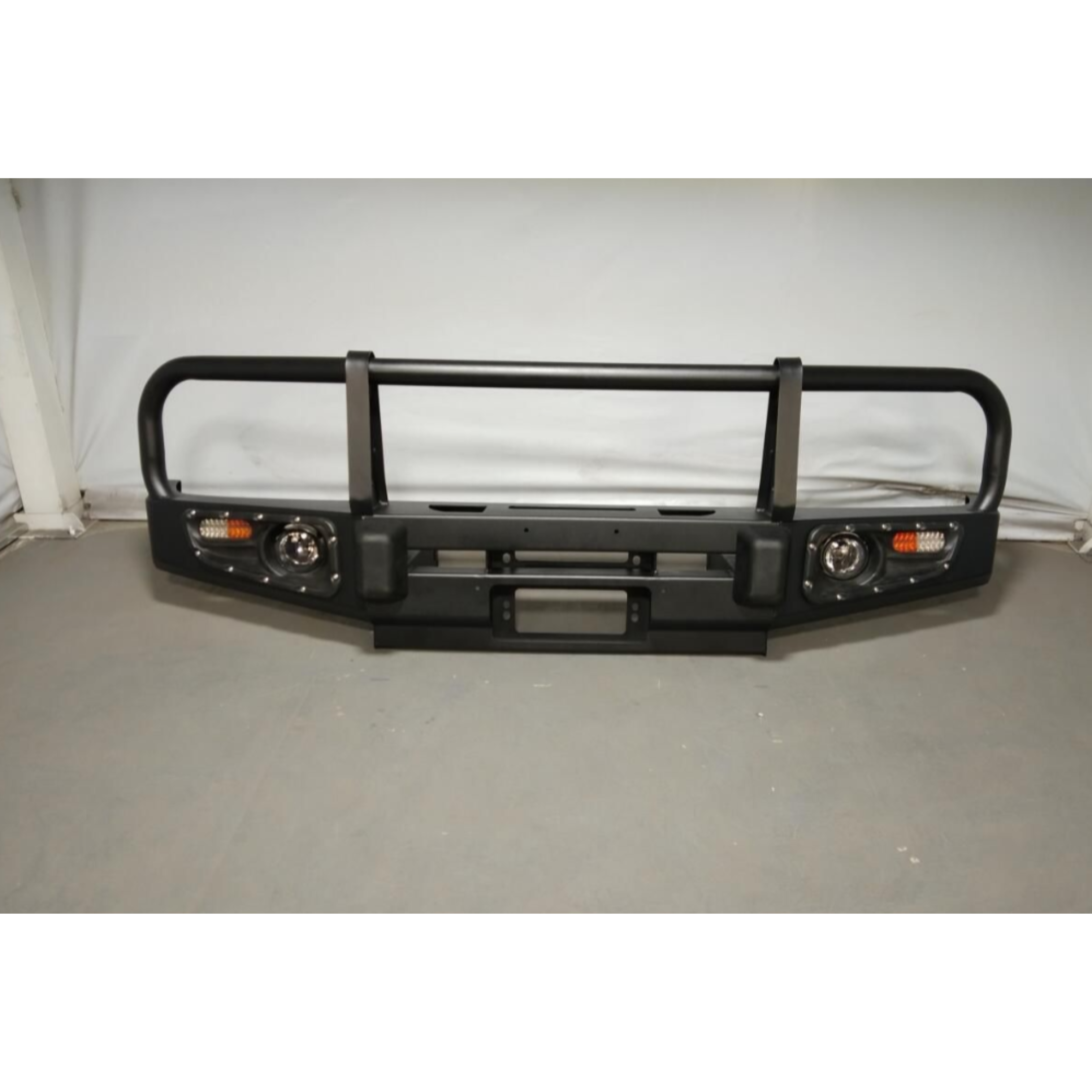 A heavy-duty, matte black steel bull bar or off-road replacement front bumper designed for a vehicle. It features a triple-hoop tubular steel design for grille and headlight protection, two integrated circular driving/fog lights, and rectangular turn signal/indicator lights with amber and white LEDs on the outer edges. The main bumper body includes mounting points and a slot for a license plate bracket. It is displayed indoors against a gray floor and a white fabric backdrop.