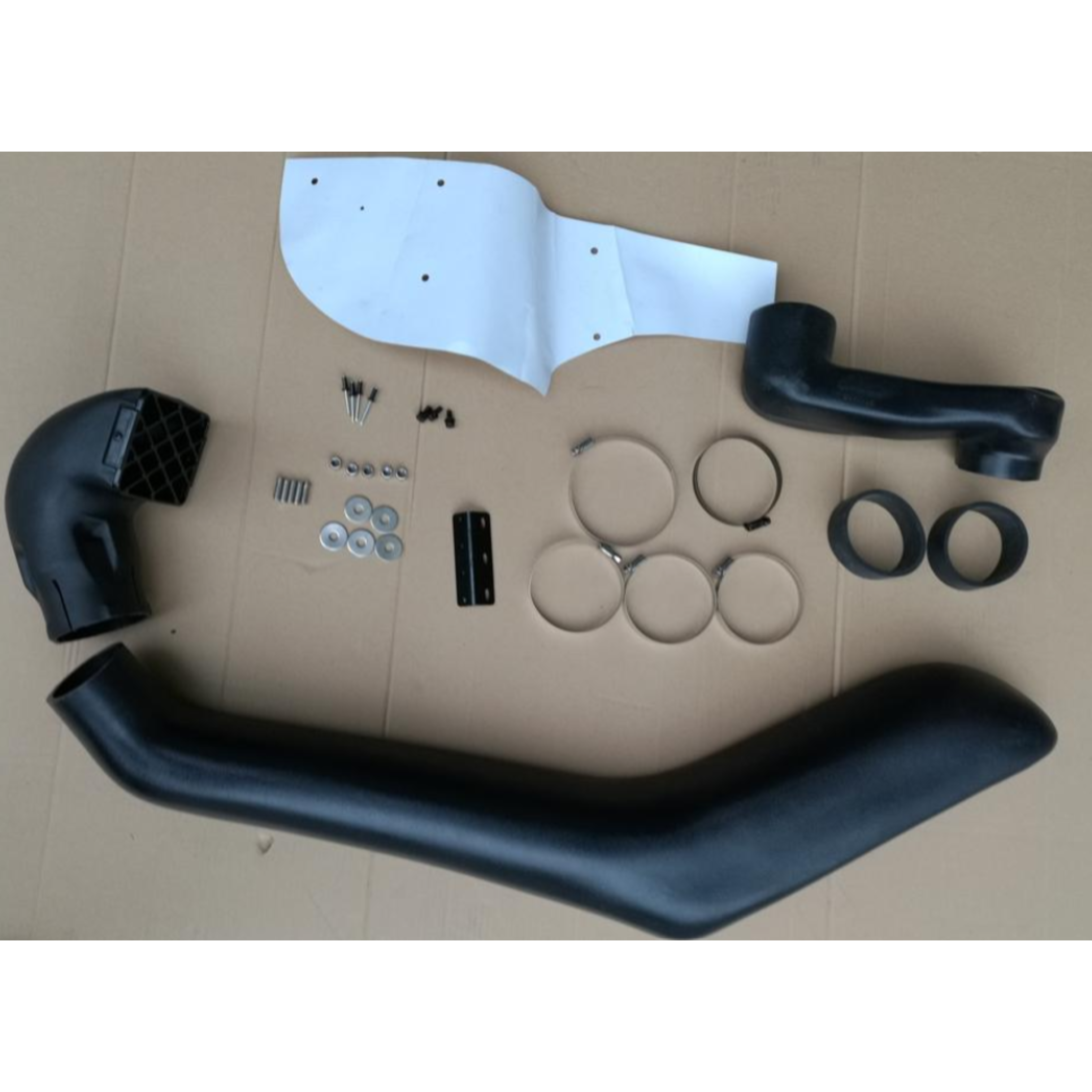 A flat lay image showing the components of a vehicle snorkel kit, mostly black plastic and metal parts, laid out on a cardboard surface.
The main components include:
A large, curved, black intake pipe (snorkel body) running along the bottom.
A black air ram/intake head with a rectangular grate on the left.
A smaller, angled black joining pipe on the upper right.
A white mounting template/plate with several holes on the top.