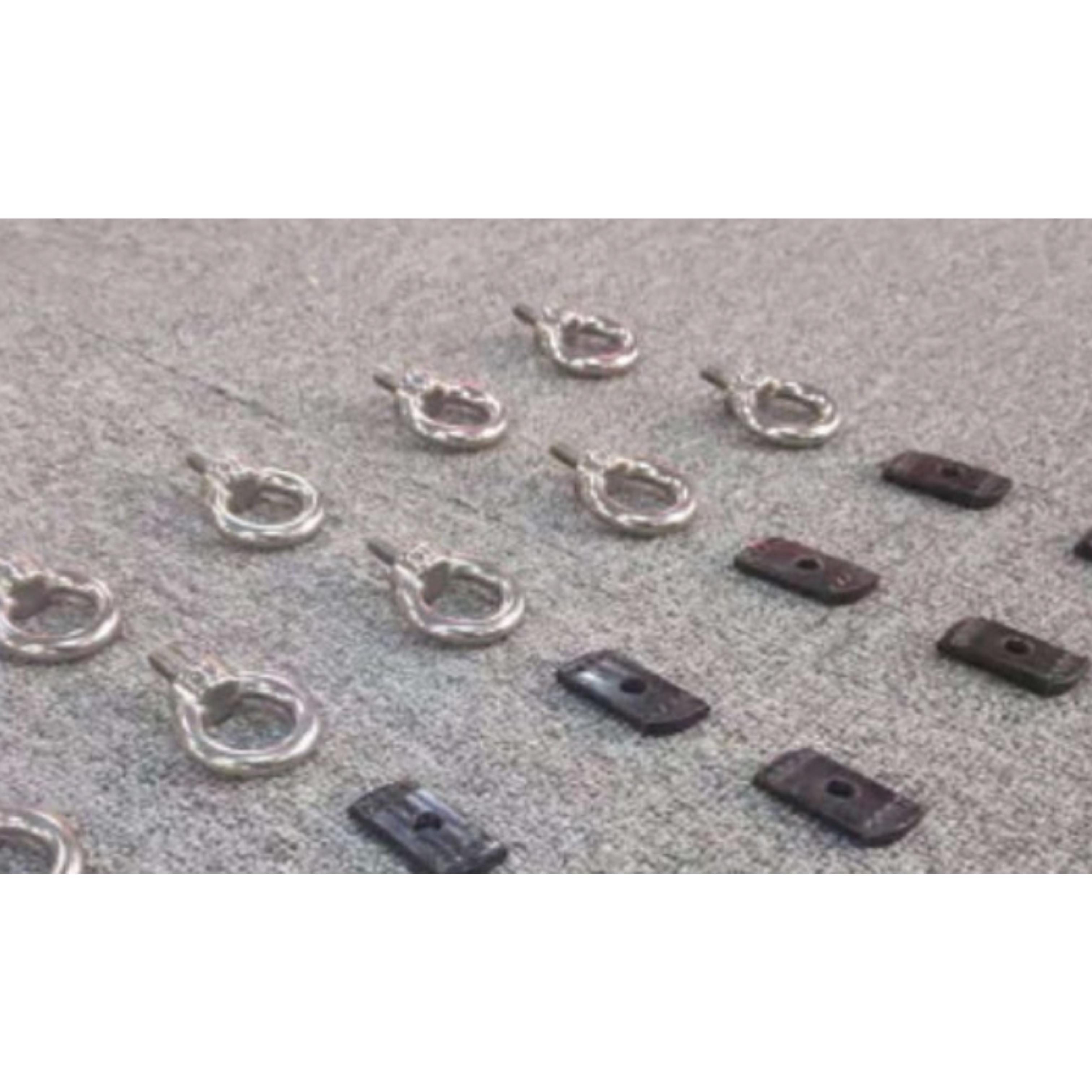 Set of chrome or stainless steel tie-down eye bolts with shanks, arranged diagonally alongside several small, rectangular black metal mounting plates, all lying on a grey, slightly coarse surface or carpet.