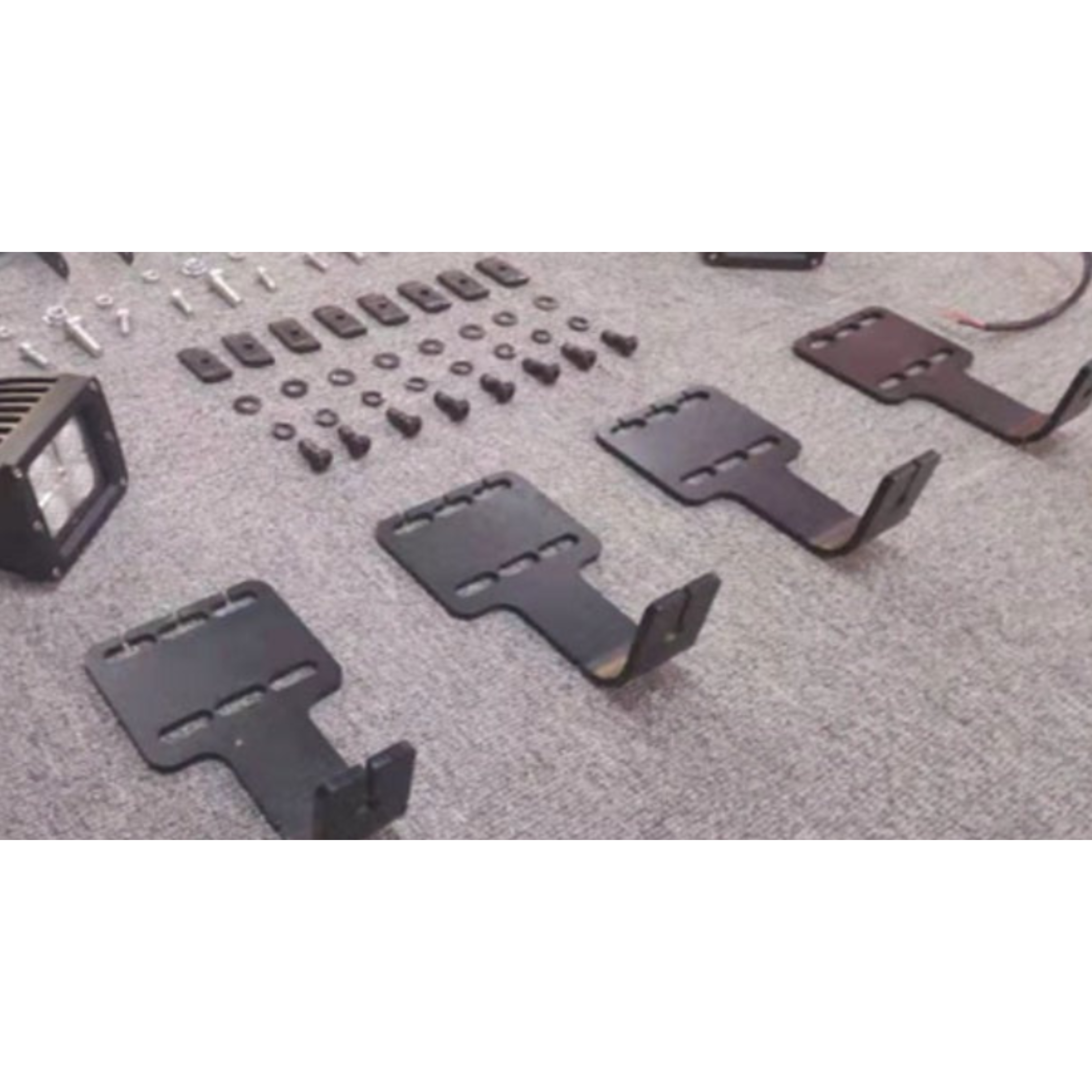 A close-up shot of automotive accessory mounting components laid out on a gray carpet. The image features four black, L-shaped metal brackets with slotted holes, arranged in the foreground. In the background, there is an assortment of black hardware (bolts, washers, and rectangular mounting plates), and a small section of a square black LED light pod is visible on the far left.