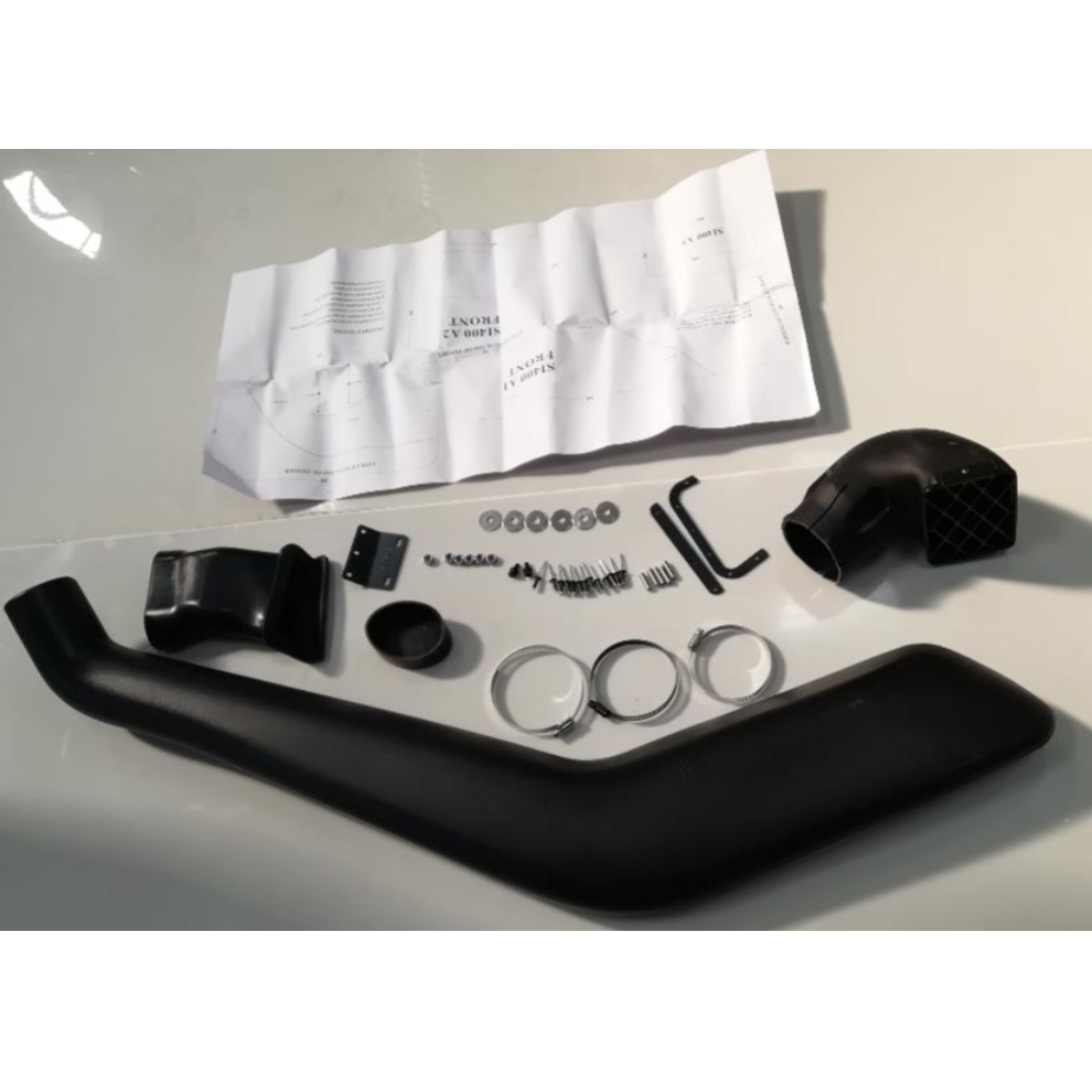 A product shot showing the components of a black vehicle snorkel kit laid out on a white surface next to a folded instruction sheet. The main component is a long, curved black plastic air intake tube. Other parts include a square-shaped air ram intake head, mounting brackets (L-shaped and flat), various nuts, bolts, washers, screws, and three large metal hose clamps. The instructions are printed on a white sheet of paper and are partially unfolded.