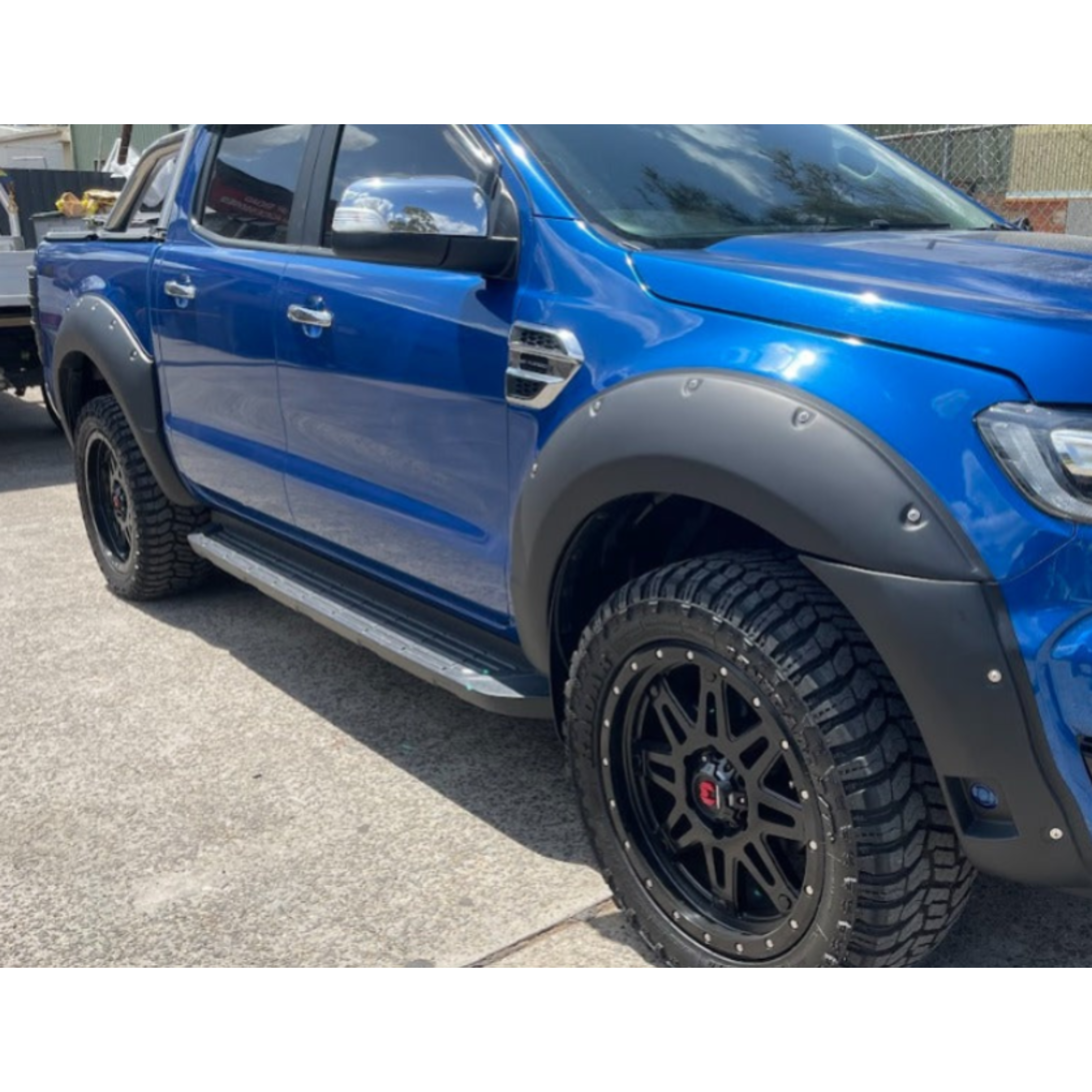 A side-angle view of a bright blue pickup truck (likely a Ford Ranger) fitted with matte black bolt-on style fender flares over all wheels. The truck is accessorized with large, black multi-spoke off-road wheels and aggressive-tread tires, and features a silver/aluminum running board or side step. The bed of the truck appears to be fitted with a tray or sports bar setup.