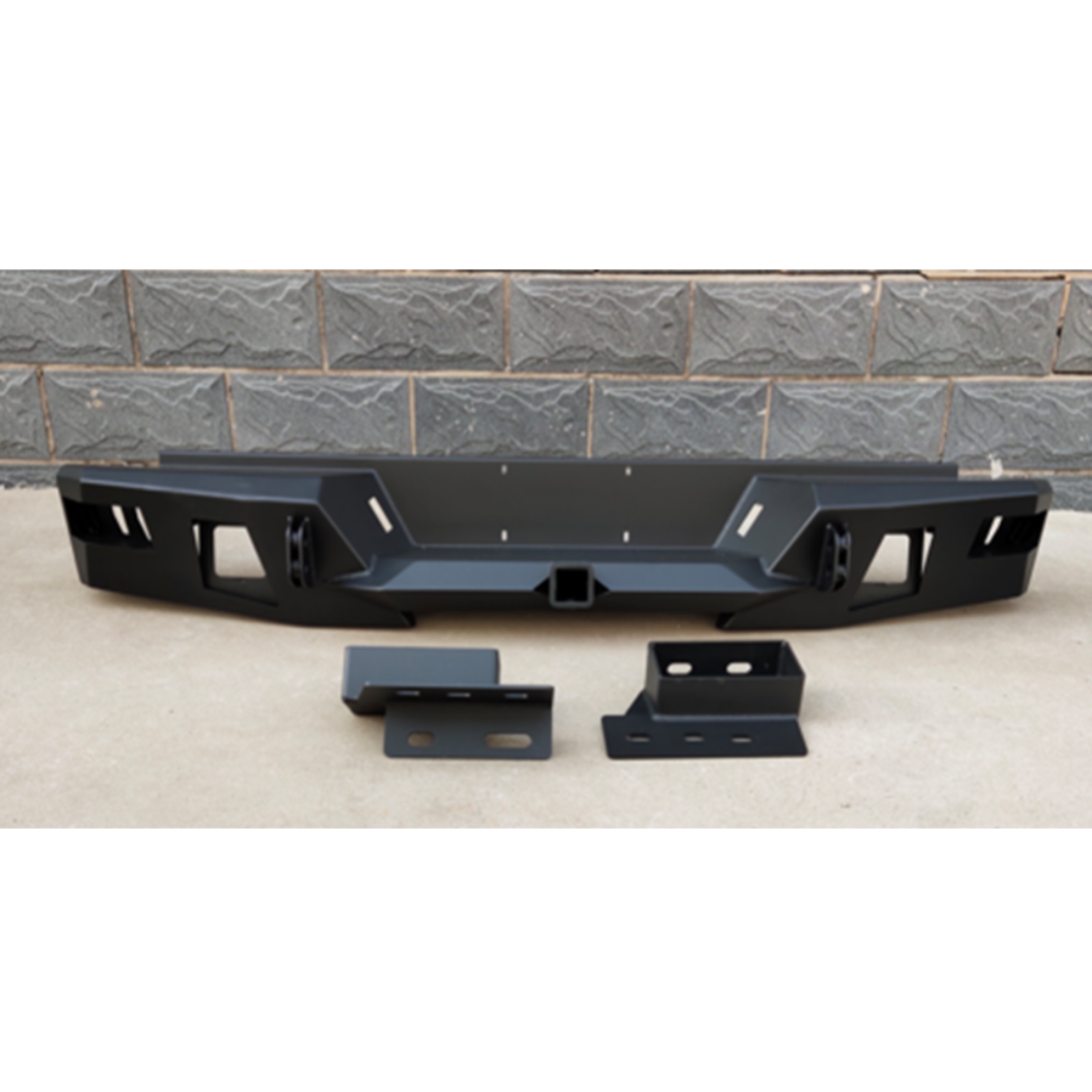 A heavy-duty, matte black steel off-road rear bumper is displayed with its mounting hardware against a brick wall background. The bumper is rugged and angular, featuring a central square receiver hitch, two prominent cutouts for D-ring shackles (one shackle is partially visible), and multiple slotted openings on the left and right ends, likely for cube lights or venting. Two large, matching black mounting brackets are placed in front of the main bumper piece.