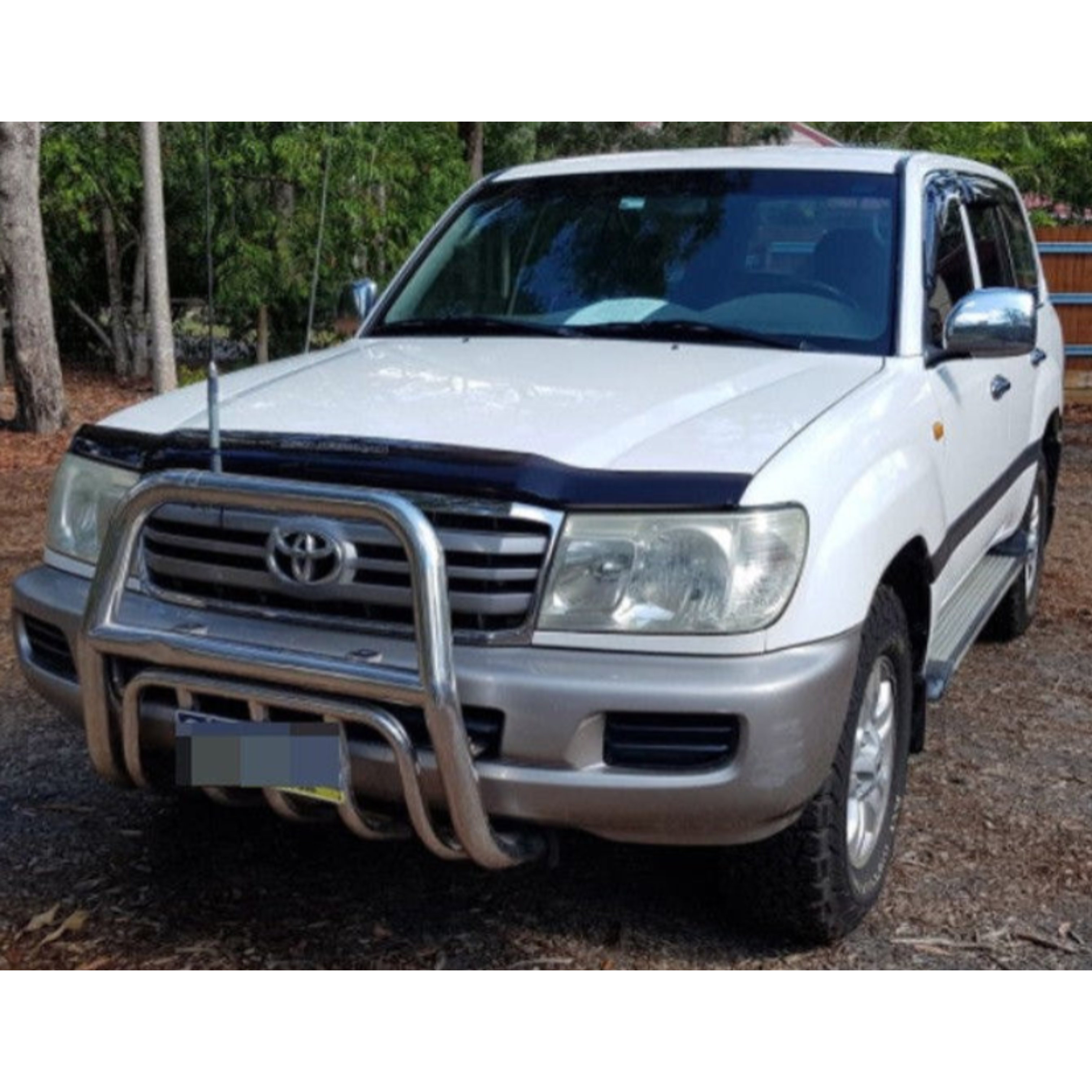 White Toyota Land Cruiser 100 Series SUV parked outdoors with a forest background. The vehicle is equipped with a chrome bull bar, a black acrylic bonnet protector on the hood, side steps, chrome mirror covers, and has gray/tan lower body trim. A vertical antenna is mounted near the driver's side front fender.