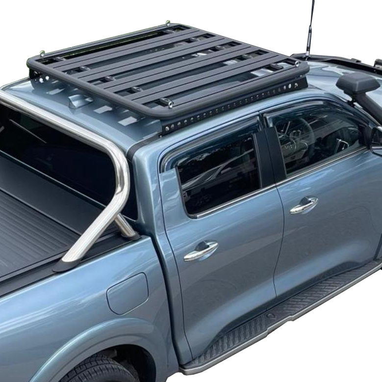 A high-angle close-up view focusing on the rear section of a blue dual cab pickup truck. The vehicle is fitted with a large flat black platform roof rack mounted above the cab. The ute tray is covered by a roller shutter (tonneau cover), and a silver or chrome sports bar is visible at the front of the tray. The truck also has side steps and dark tinted windows. The image is isolated against a white background.