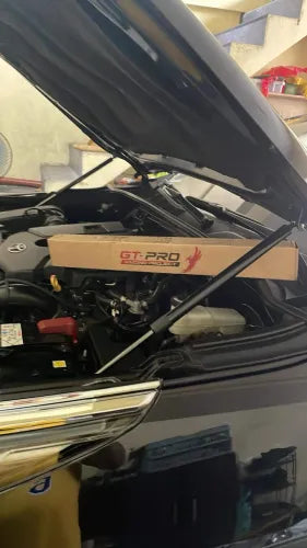 A dark-colored car (likely black) is shown with its hood open, revealing the engine bay. A pair of hydraulic hood struts are installed and holding the hood up. A long, brown cardboard box with the "GT-PRO" logo is resting horizontally across the engine area, suggesting it is the packaging for the newly installed struts. The car is parked indoors near a set of stairs.