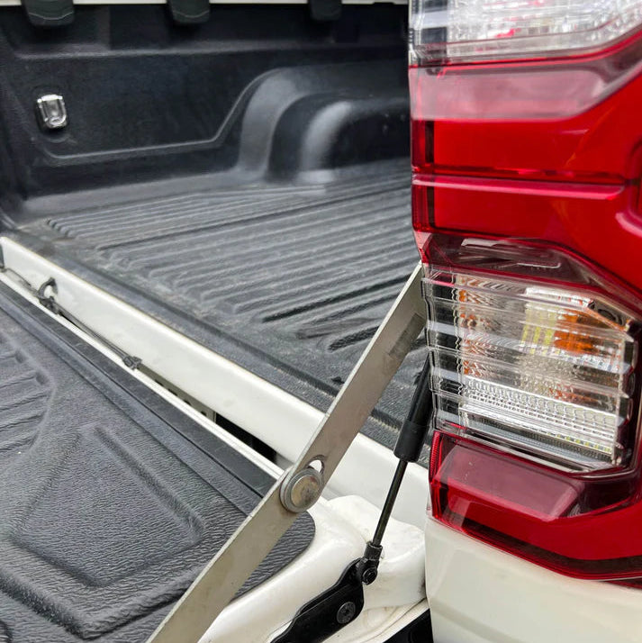A close-up photograph focused on the tailgate of a white pickup truck, which is open. The image highlights a tailgate assist strut installation. A black hydraulic damper rod is visible, connected to a silver metal linkage/limiting arm. The truck bed has a black textured bed liner, and the red and clear taillight assembly is prominent on the right side.