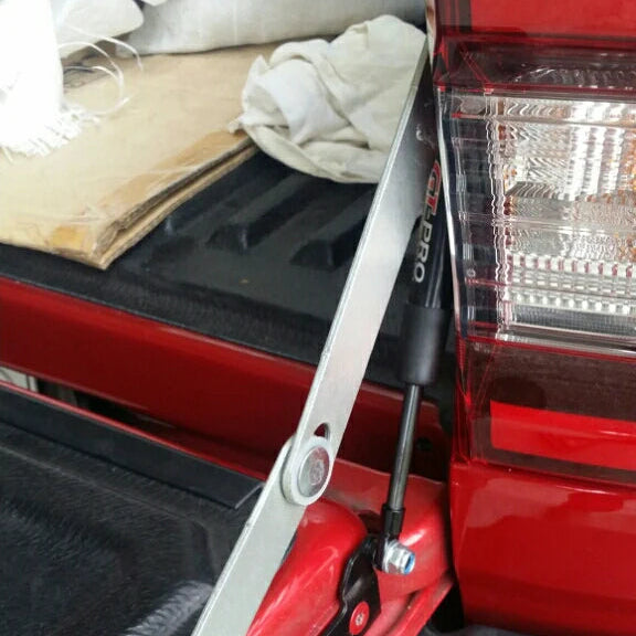 A close-up image showing an installed tailgate damper or assist strut on a red pickup truck. A black hydraulic strut labeled "GT-PRO" is visible, connecting a silver metal bracket to the tailgate/bed. The bracket is bolted near the edge of the tailgate, which has a textured black bed liner surface. The truck's red taillight assembly is visible on the right.
