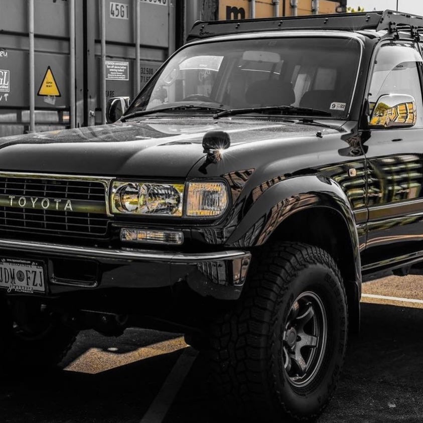 A low-angle, black and white photograph of the front corner of a black Toyota Land Cruiser (likely an FJ80 series), parked outdoors. The vehicle has large, off-road tires on aftermarket wheels, flared fender arches, a roof rack, and a prominent "TOYOTA" grille badge. The background features a large cargo container with markings.