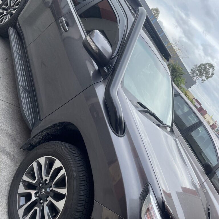 A slightly low-angle, close-up shot of a dark grey Toyota Land Cruiser Prado, showing the front passenger side. A black snorkel is prominently installed, running up the A-pillar and connecting to the engine's air intake near the hood. The vehicle also has a black side step, a multi-spoke alloy wheel with black accents, and a portion of a roof rack visible. The image is taken outdoors on a cloudy day, with what appears to be a dealership or parking lot background.