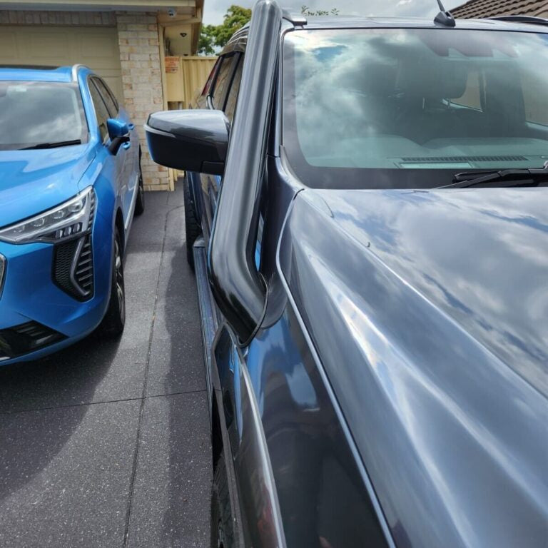 A slightly low-angle, close-up shot focusing on the front passenger side of a dark grey SUV or pickup truck, parked next to a bright blue SUV (possibly a Haval). A glossy black snorkel is prominently installed, running up the A-pillar. The vehicle's hood reflects a cloudy blue sky. Both vehicles are parked on a dark tiled driveway, with a beige brick garage and fence visible in the background.