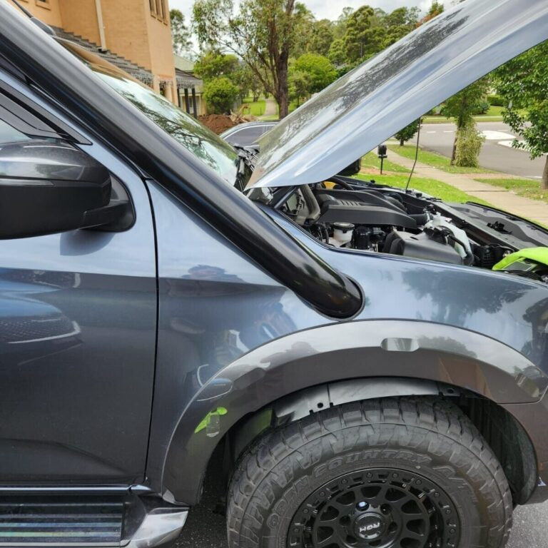 A close-up side view of a dark grey SUV or pickup truck parked outdoors in a suburban setting. The hood is open, revealing the engine bay. A glossy black snorkel is installed on the passenger side fender. The vehicle is fitted with black aftermarket fender flares and a knobby all-terrain tire mounted on a black multi-spoke wheel. A silver side step is also visible. The background shows houses, a paved street, and trees under an overcast sky.