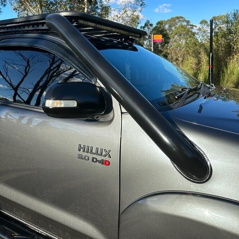 A close-up side view of a grey Toyota Hilux 3.0 D-4D with a large, black, aftermarket snorkel attached to the A-pillar and extending up along the windshield. The vehicle also has a black roof rack with an LED light bar mounted on the front edge and a tall antenna visible on the passenger side. The vehicle is parked outdoors with trees and a blue sky in the background.