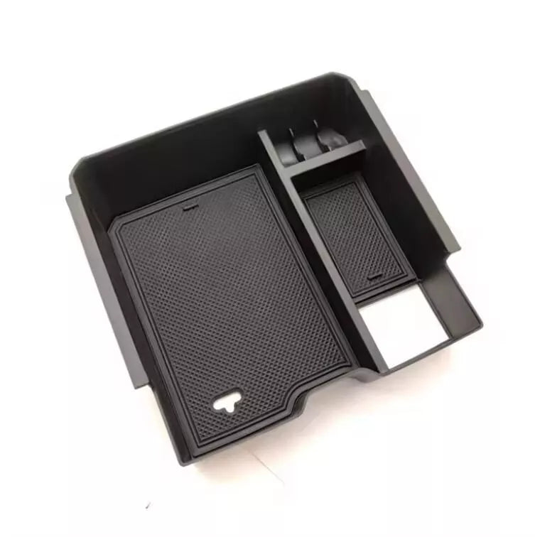 A black plastic car center console organizer tray insert, photographed from a slight angle against a white background. The tray is rectangular and features multiple compartments.
The largest section on the left contains a removable, non-slip rubber mat with a textured, hexagonal pattern and a small U-shaped cutout at the bottom. The right side of the tray is divided into a smaller rectangular section, also with a textured rubber mat, and a small, two-slot holder at the back. A square opening or cutout is