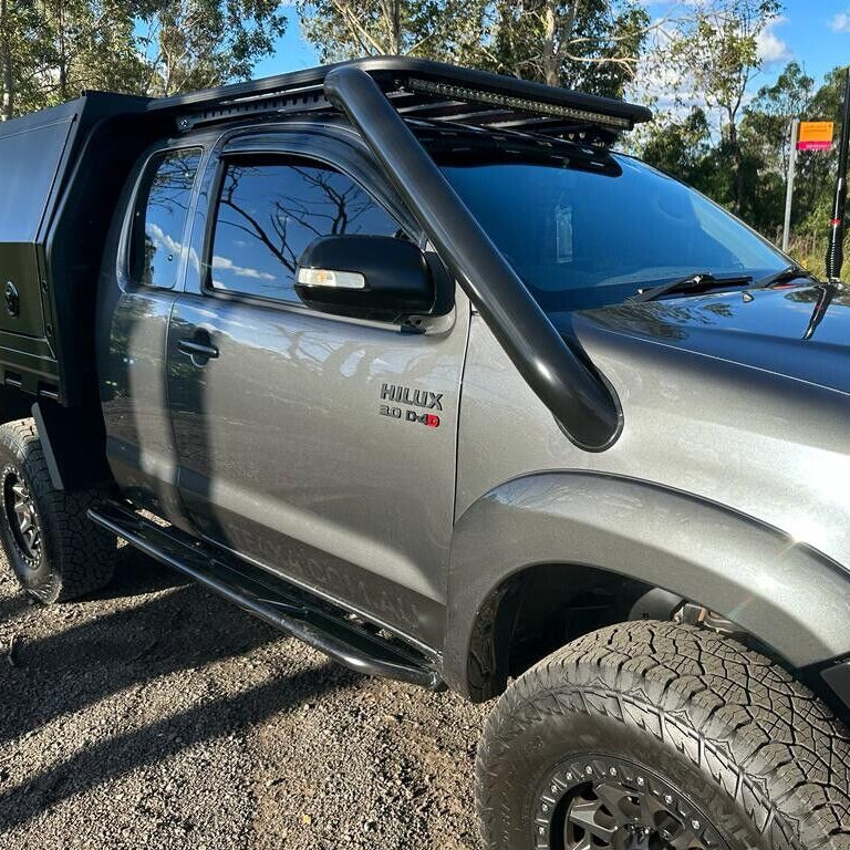 A detailed side view of a grey Toyota Hilux 3.0 D-4D dual cab ute heavily modified for off-roading. Key features visible include a large, black snorkel, black rock sliders/side steps, a black roof rack with an LED light bar, and all-terrain tires mounted on black aftermarket wheels. The rear of the truck has been converted to a black utility tray/canopy. The vehicle is parked on gravel under a blue sky with trees in the background.