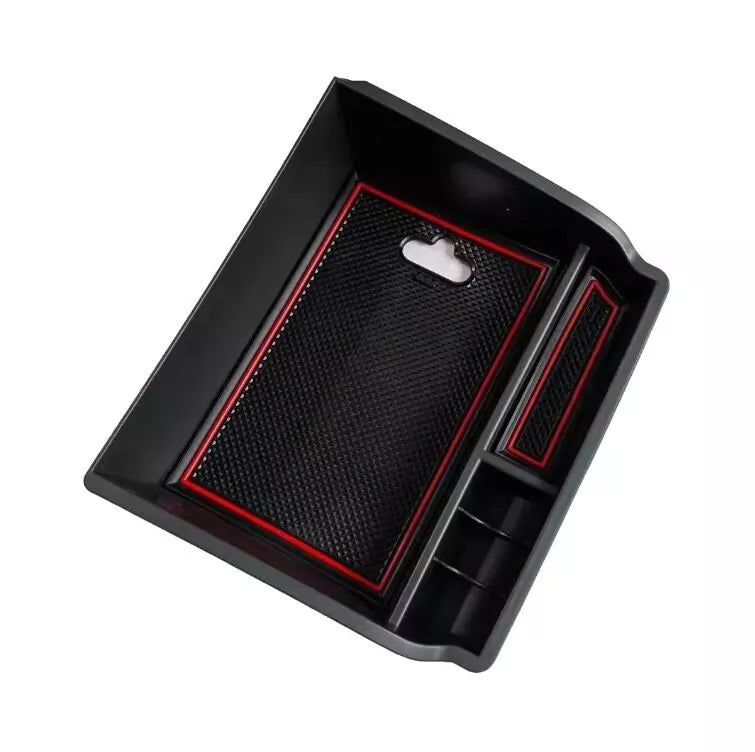 A black plastic storage insert designed to fit into a vehicle's armrest or console. It features a large main compartment and smaller side sections, with a removable black rubber mat and red accent trim lining the sections.
