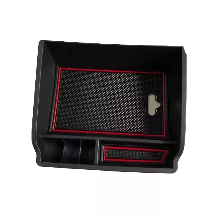 A high-angle product image of a black plastic center console organizer tray for a vehicle, isolated on a white background. The tray is rectangular and features multiple storage compartments. It includes removable black anti-slip rubber mats with a diamond pattern, which are accented with a bright red border around the edges.